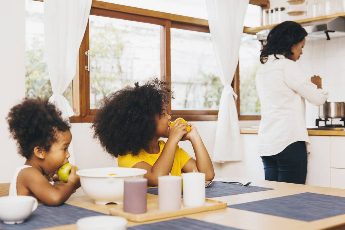 Two young girls snack at the kitchen table while their mother works at the stove.