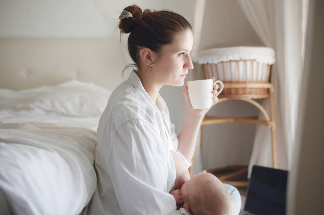 Tired mum, holding a coffee mug while nursing a newborn
