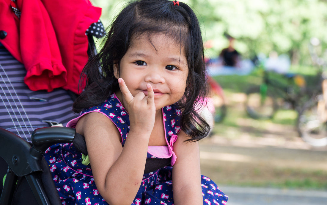 Toddler girl in a stroller at a park using sign language to say Love