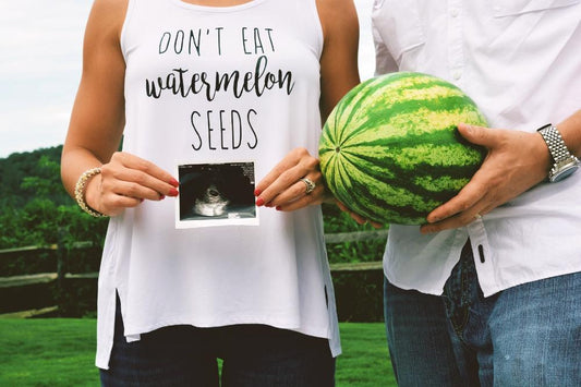Woman holding ultrasound wearing shirt that says "don't eat watermelon seeds" standing next to a man holding a watermelon