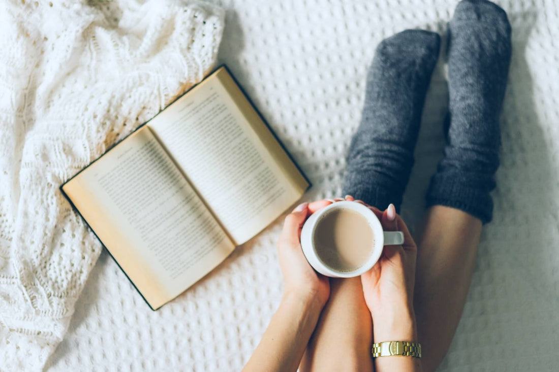 A woman lounges next to an open book in cosy socks as she holds a cup of coffee