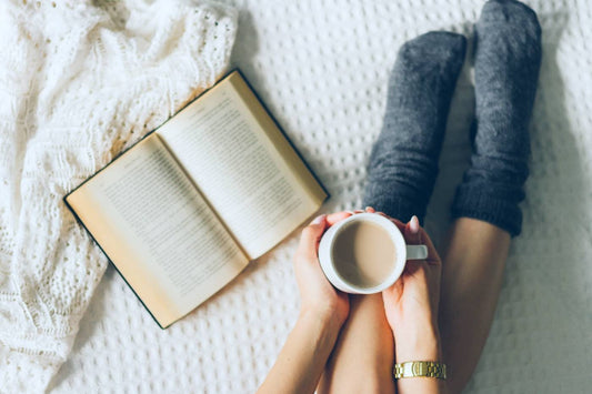 A woman lounges next to an open book in cosy socks as she holds a cup of coffee