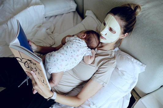 postpartum self-care: a new mom wearing a face mask and reading while a baby sleeps on her chest