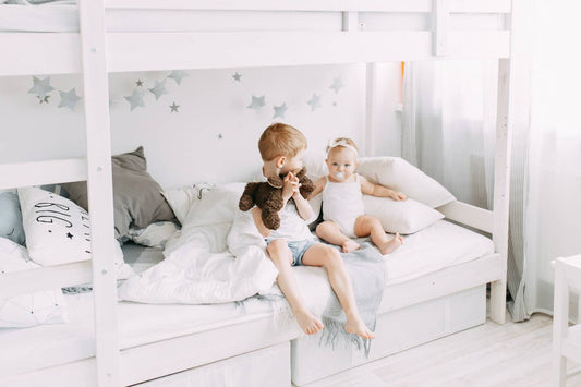 A young boy and his toddler sister sit on the bottom of a bunk bed.