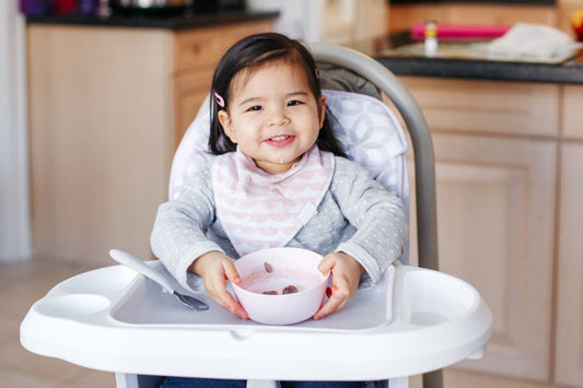 Toddler girl smiles as she sits in a highchair and holds a bowl of food